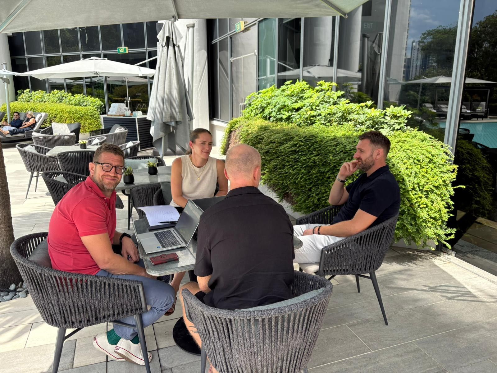 Three people sit at a table on an outdoor patio, one using a laptop while engaged in a professional discussion.