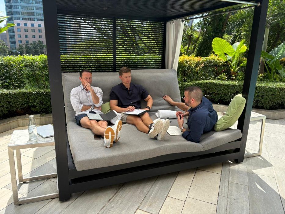 Three men sit on a large outdoor lounge chair in a covered cabana, reviewing documents together in a garden setting.