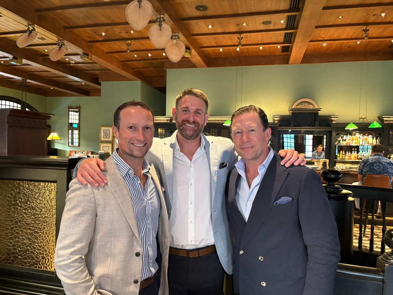 Three men smiling together in a restaurant with warm ambient lighting and a bar in the background.