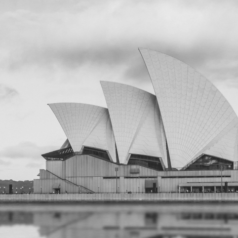 The Sydney Opera House featuring its iconic white sail-shaped roofs against a cloudy sky, in monochrome.