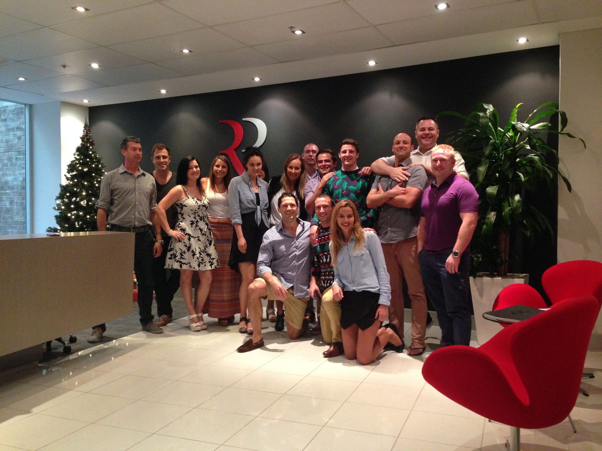 A diverse group of colleagues pose together for a photo in a modern office lobby with festive decor and red chairs.