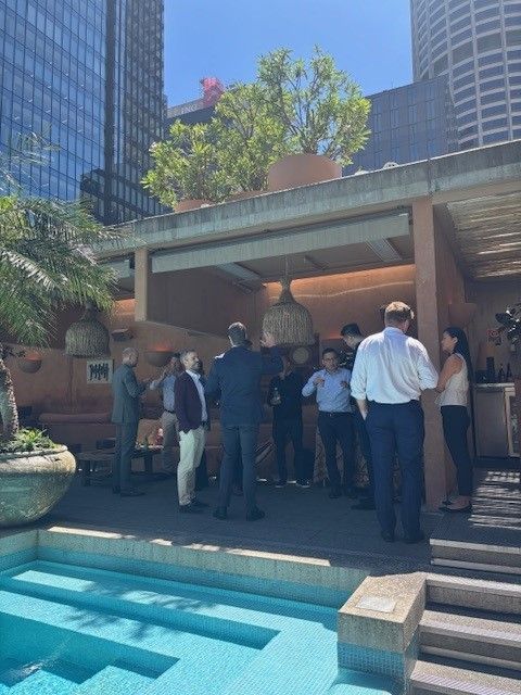 A group of people in business casual attire socialize outdoors on a rooftop terrace near a pool, framed by tall buildings.