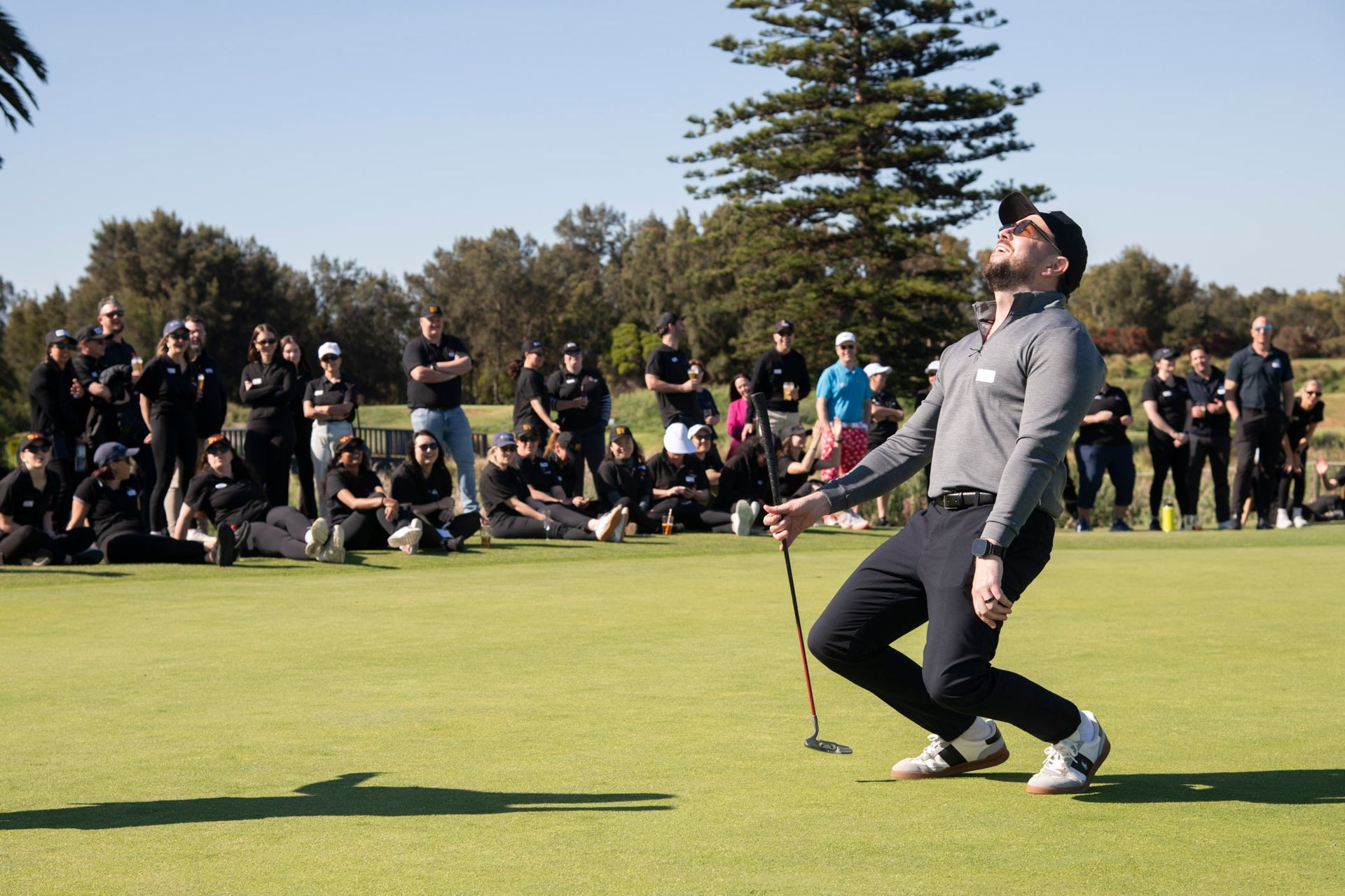 A person on a golf green reacts with emotion, holding a club while a crowd watches from the background on a sunny day.