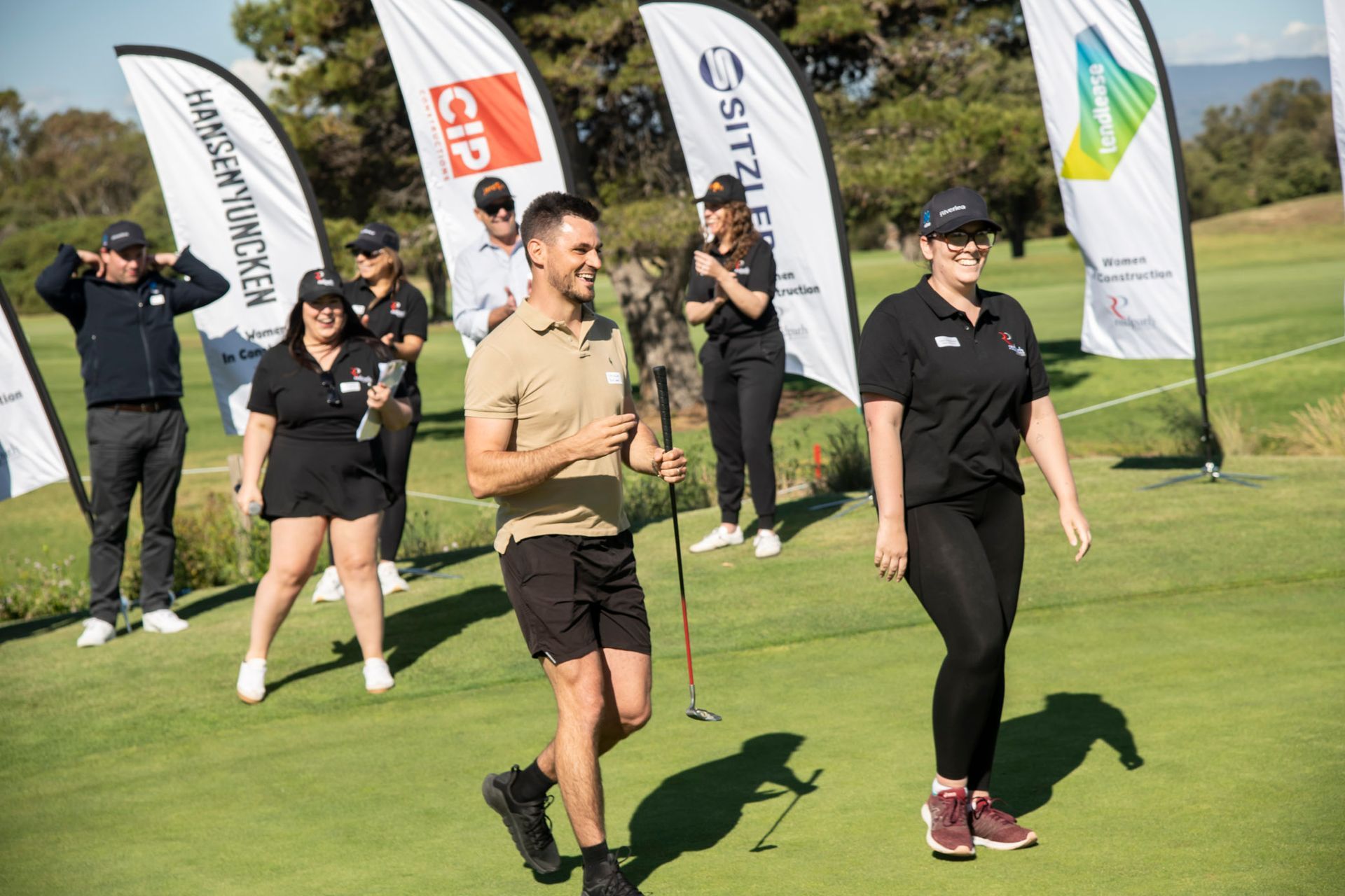 People walking and smiling on a golf course in front of sponsor flags during a sunny outdoor event.