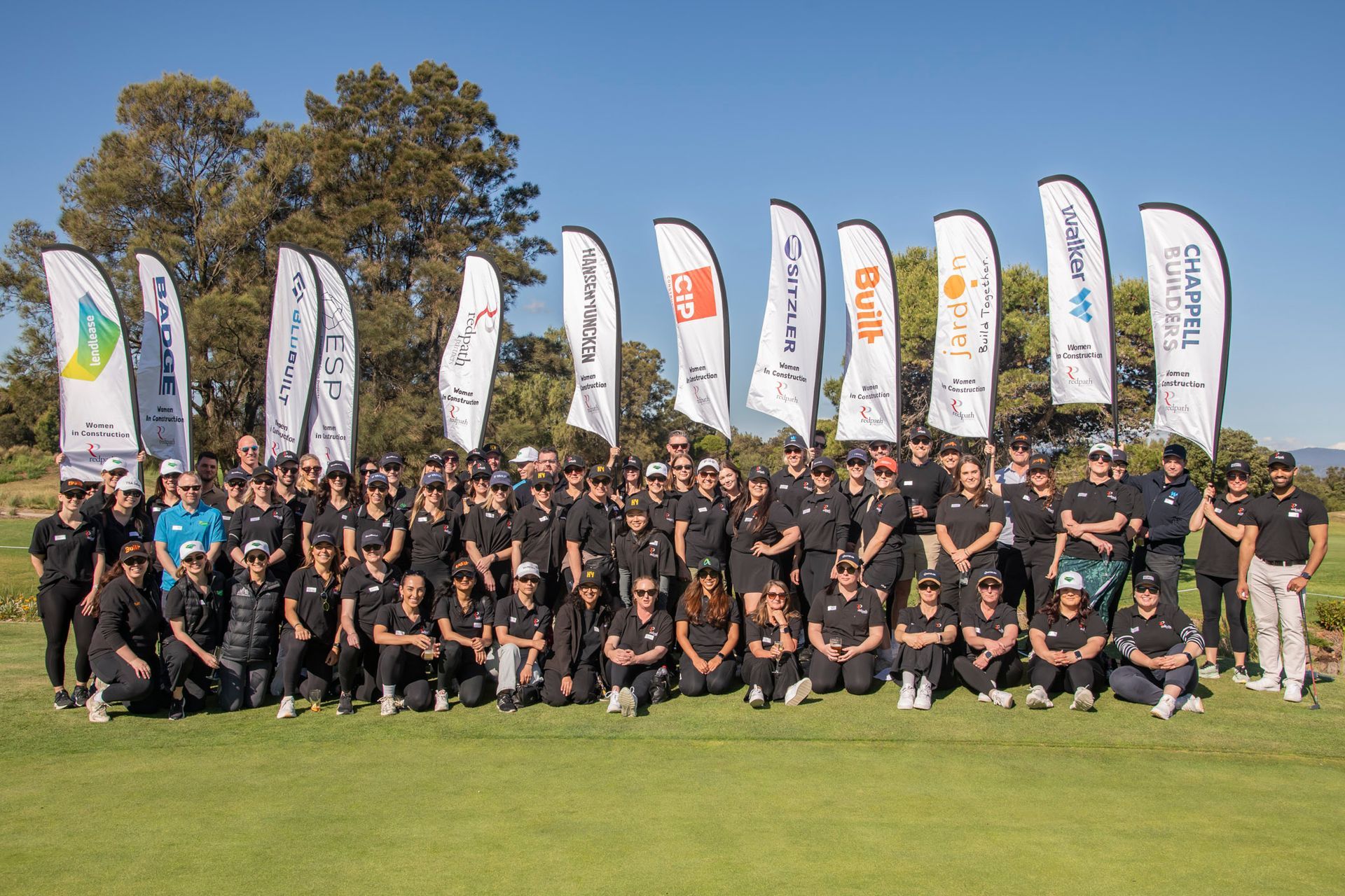 A large group of people in matching black shirts pose together on a sunny golf course in front of several sponsor flags.