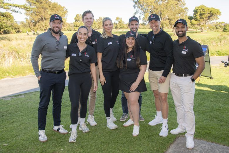 A group of eight people wearing branded shirts and hats posing together on a grassy field outdoors.