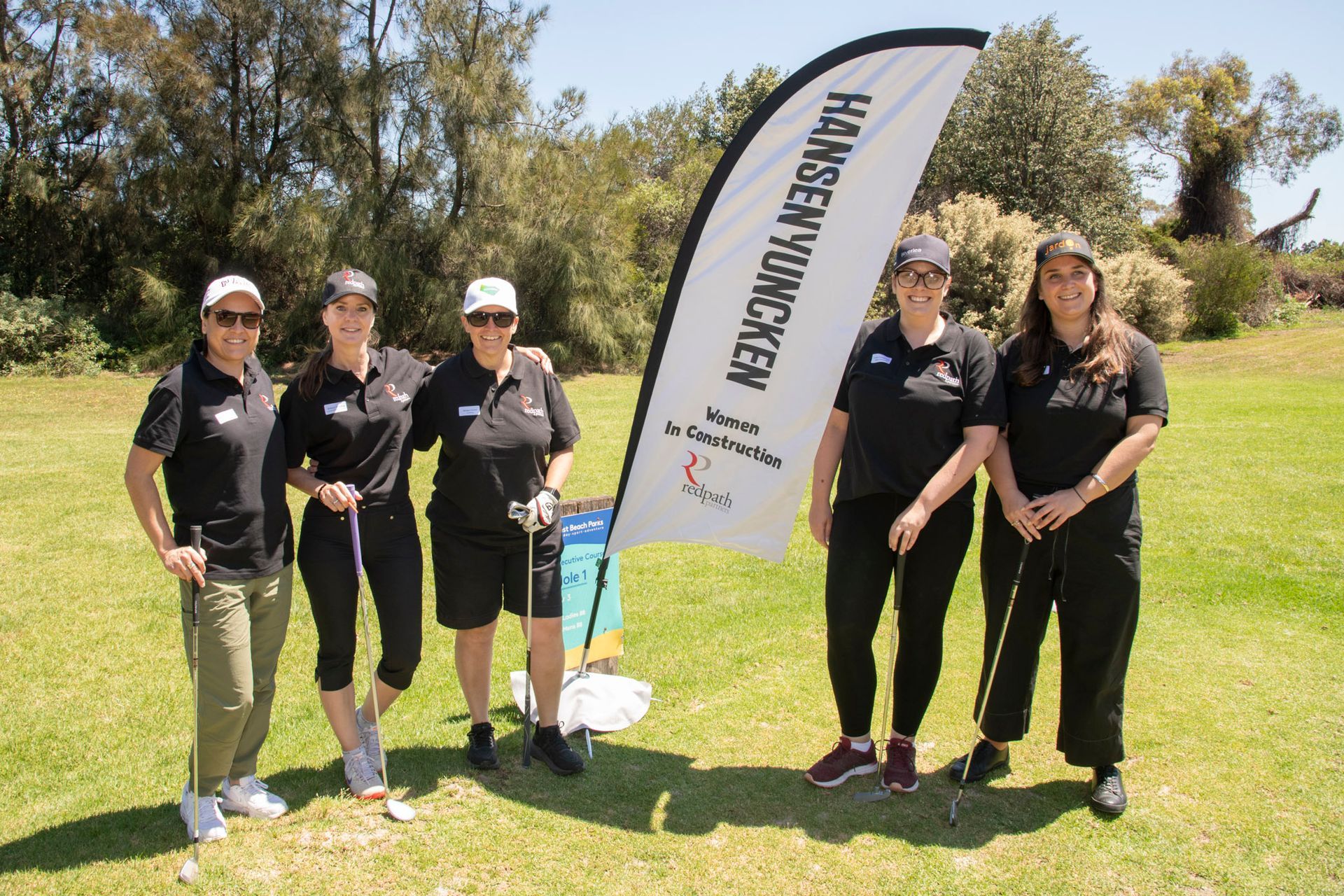 Five people wearing black shirts and caps pose on a grassy field next to a Hansen Yuncken branded banner.
