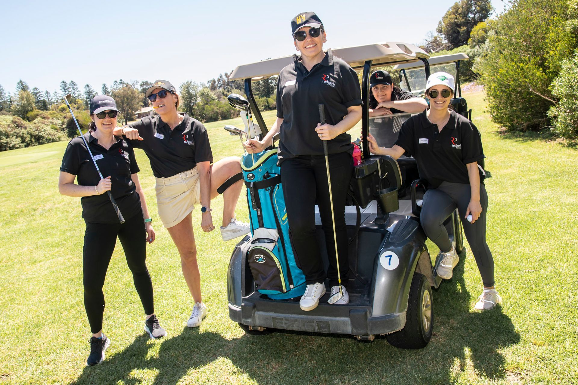 Five people in matching black shirts pose together by a golf cart on a sunny, grassy golf course.