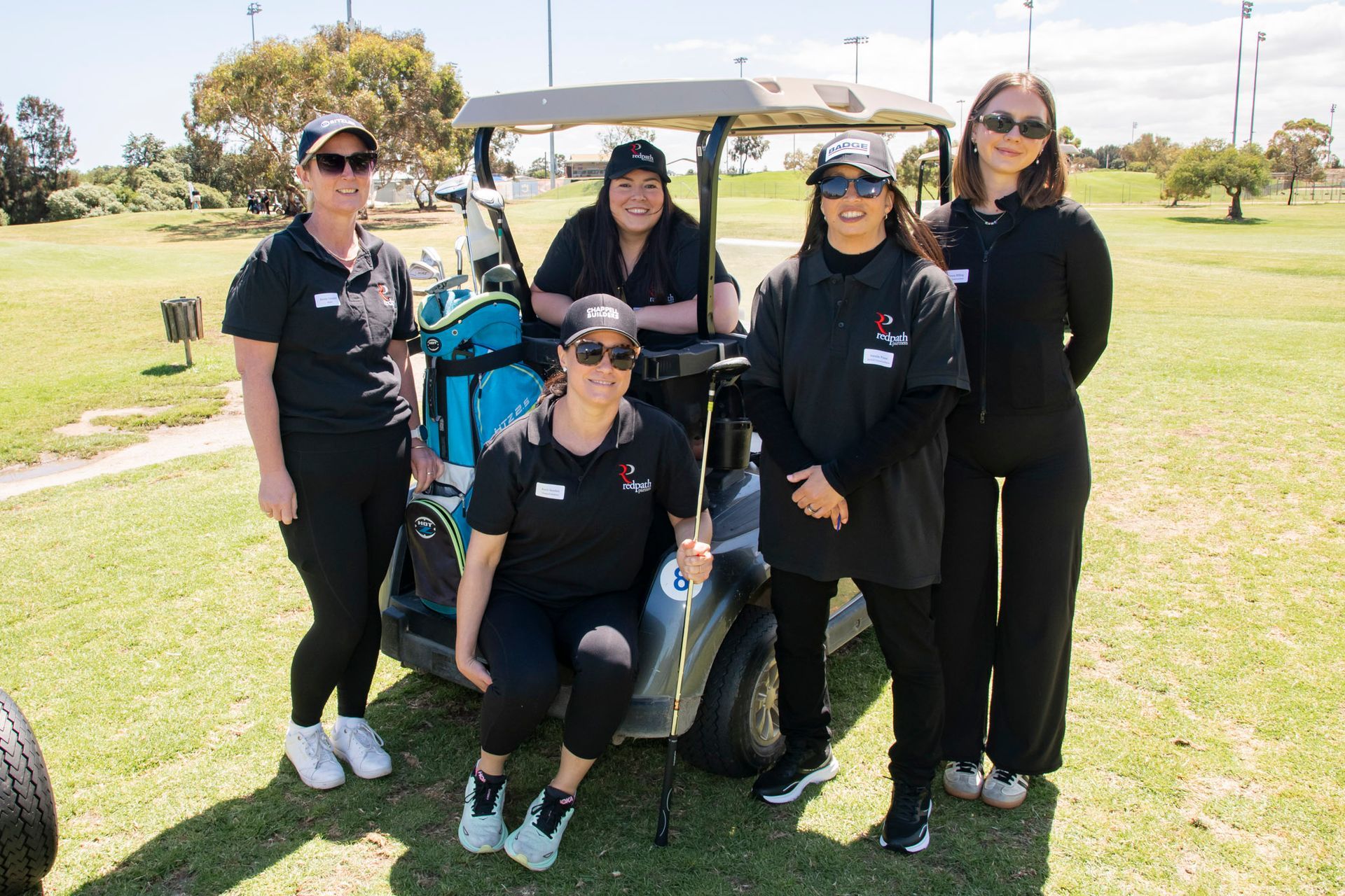 Four people in black shirts and hats pose smiling next to a golf cart on a sunny, grassy golf course.