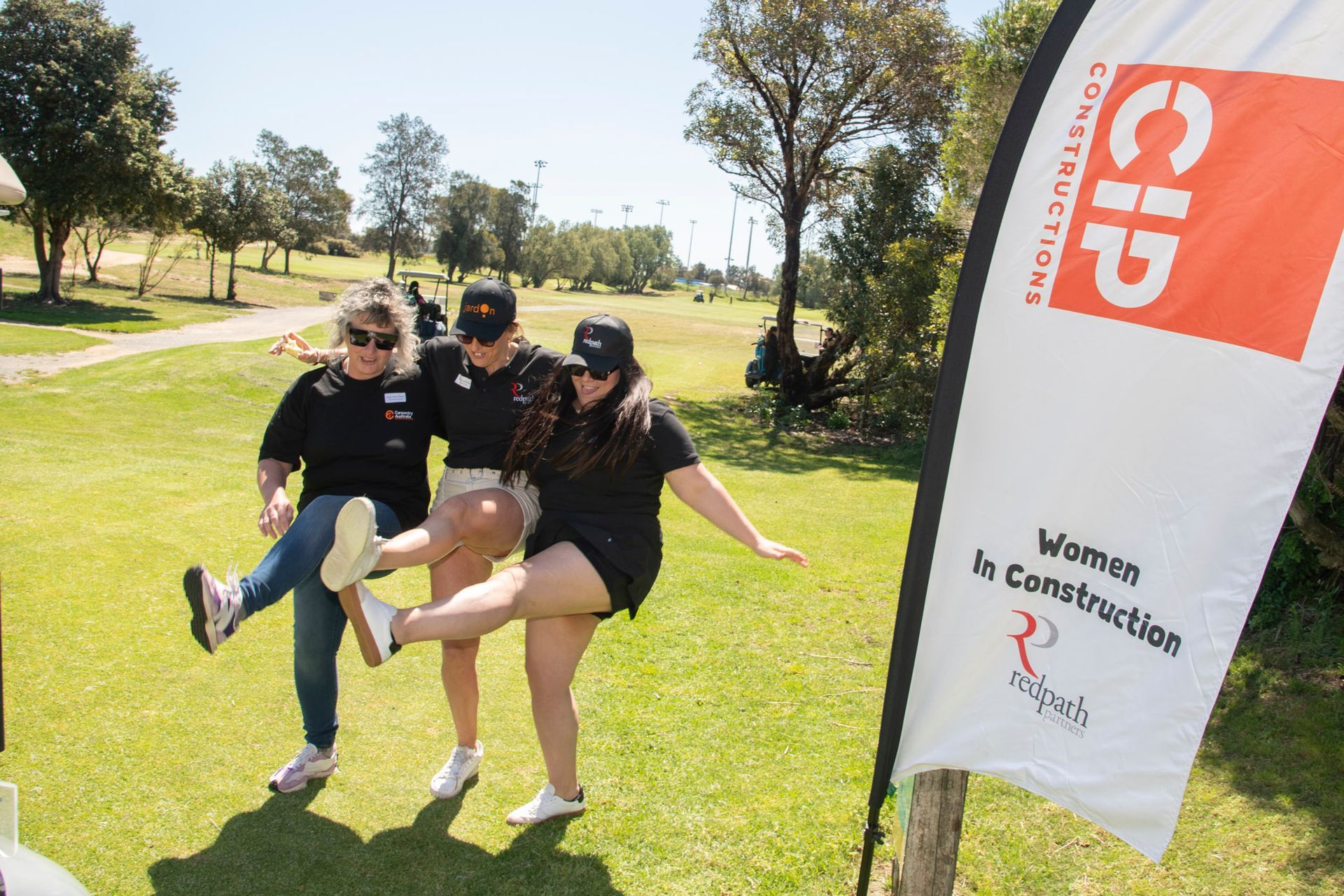 Three women posing and kicking their legs together on a sunny golf course next to a Women in Construction banner.