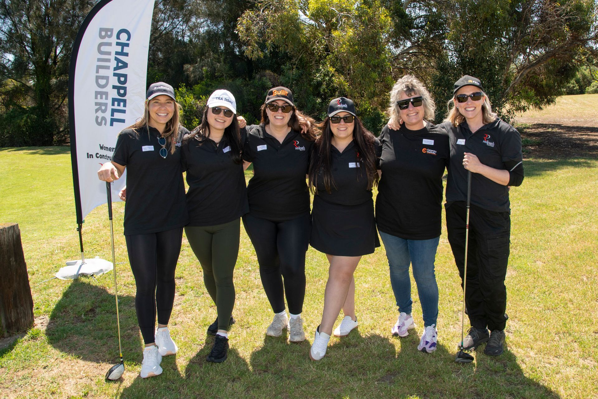 Six smiling people stand in a row outdoors in matching black shirts at a golf course, next to a Chappell Builders banner.