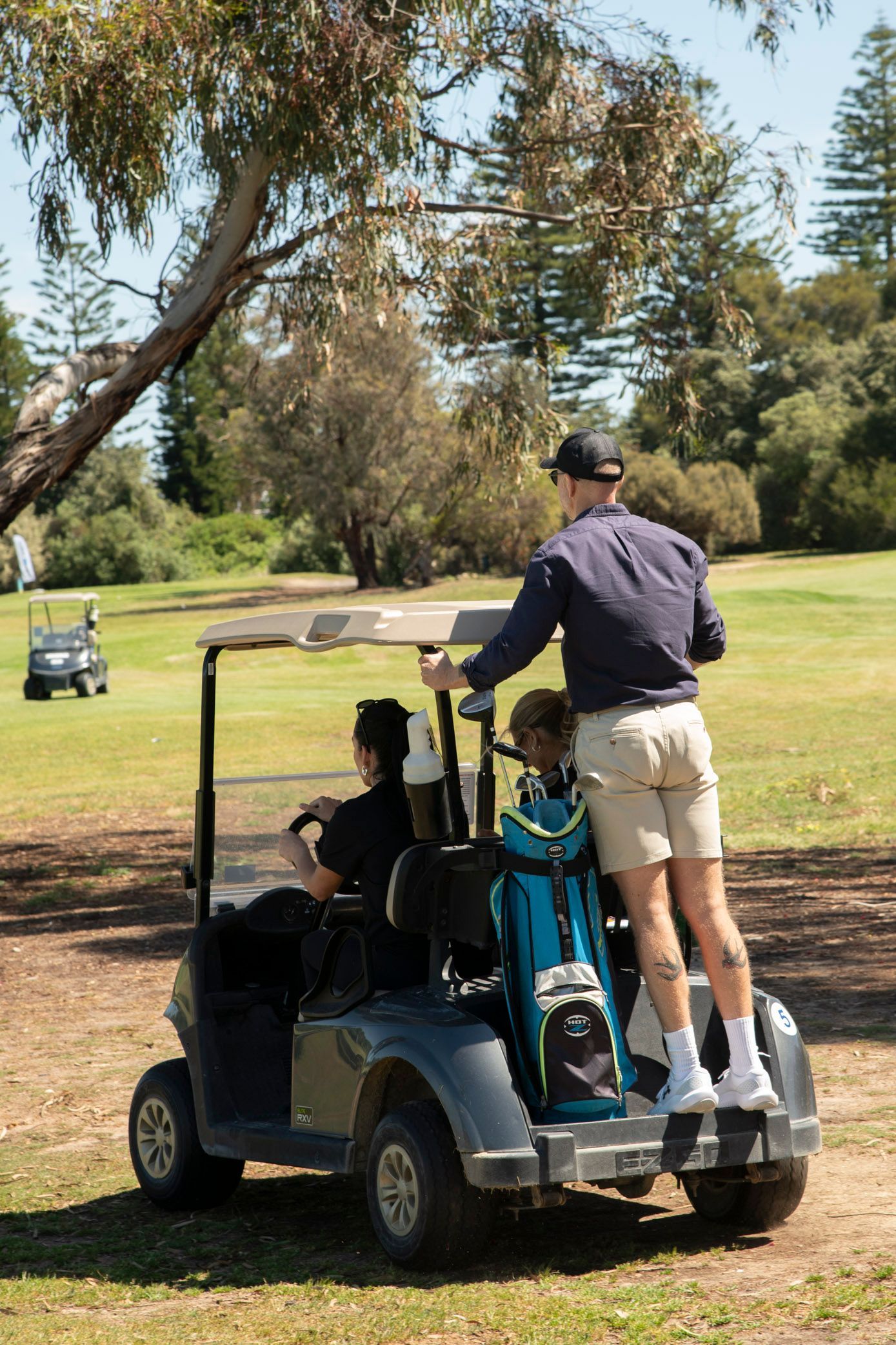 A person stands on the back of a golf cart driven by another person through a sunny, tree-lined golf course.