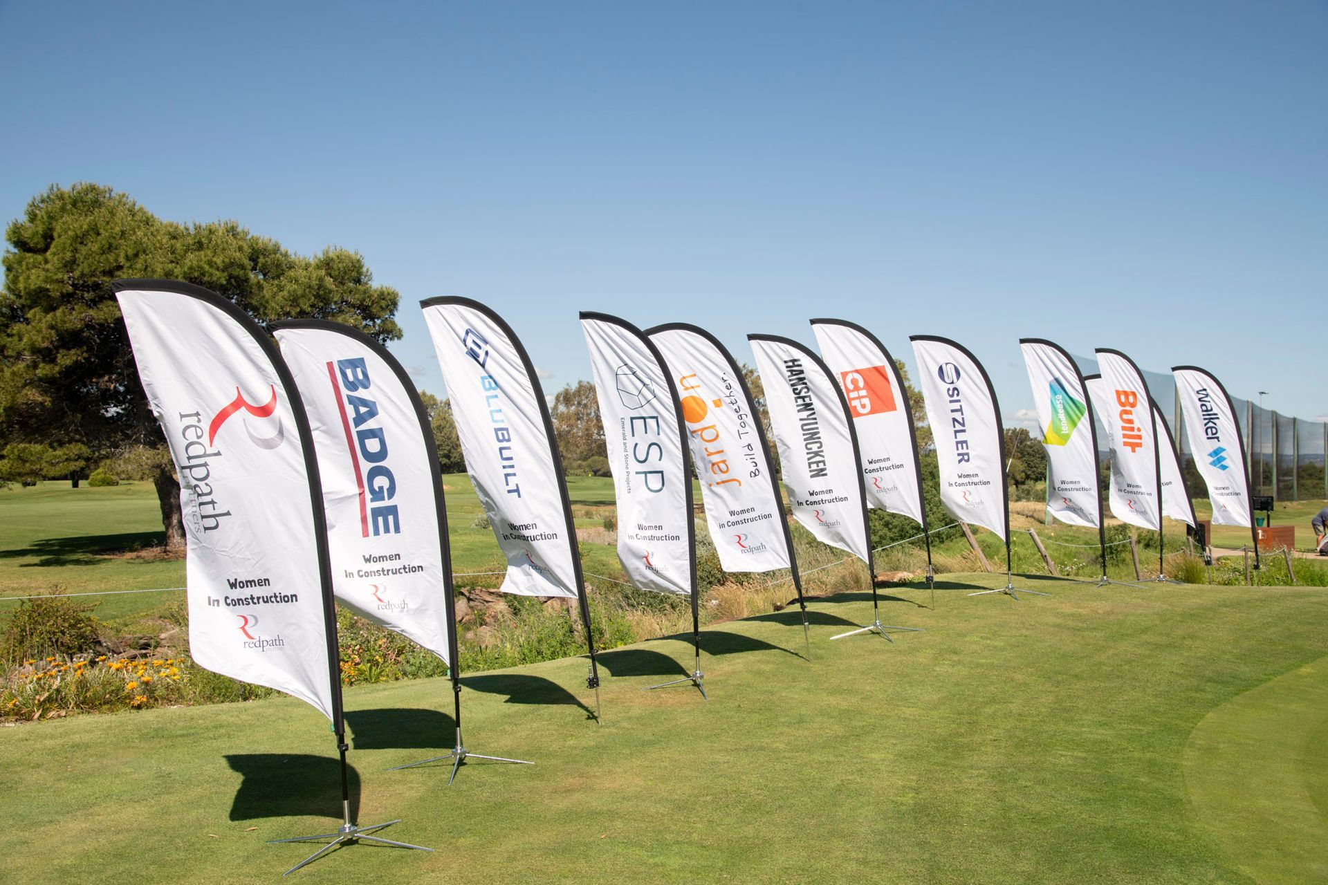 A row of white, branded feather flags standing on a grass golf course under a clear blue sky.