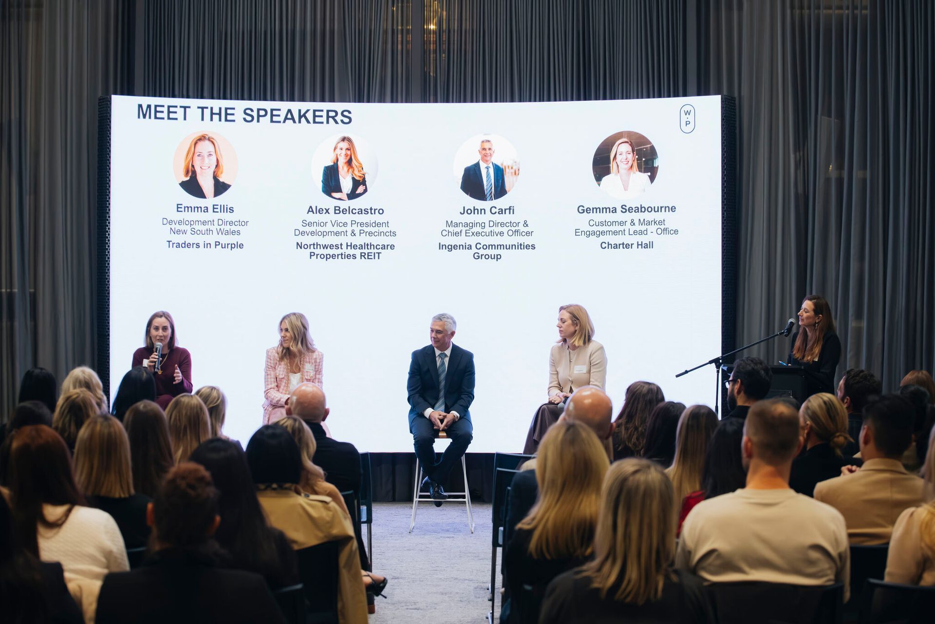 A panel of four speakers sits on a stage before an audience, with a digital display listing their names and titles above.