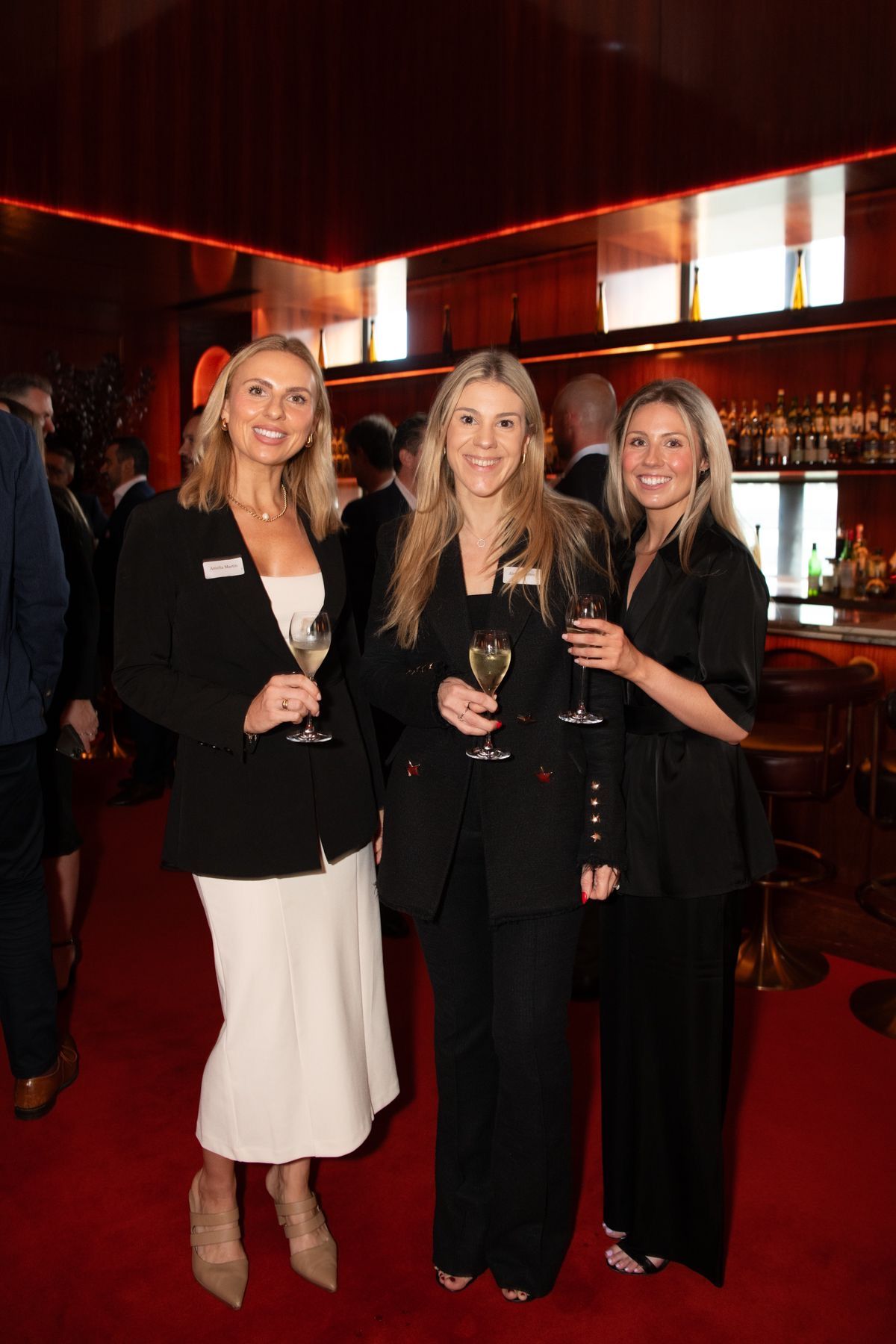 Three individuals in professional attire smile while holding drinks at an evening event with a red carpet and bar.