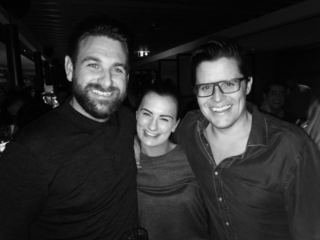 Three smiling people stand close together for a photo in a dimly lit indoor setting.