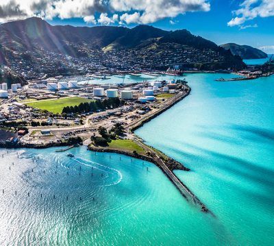Aerial view of a coastal industrial harbor with bright turquoise water, white storage tanks, and hills in the background.