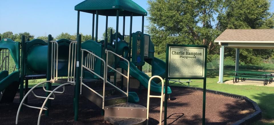 Playground with green climbing structure and ramp beside a park sign, under a pavilion on a sunny day