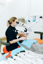 Dentist examining a patient's mouth with a microscope. The patient is lying down. Dental equipment is visible.
