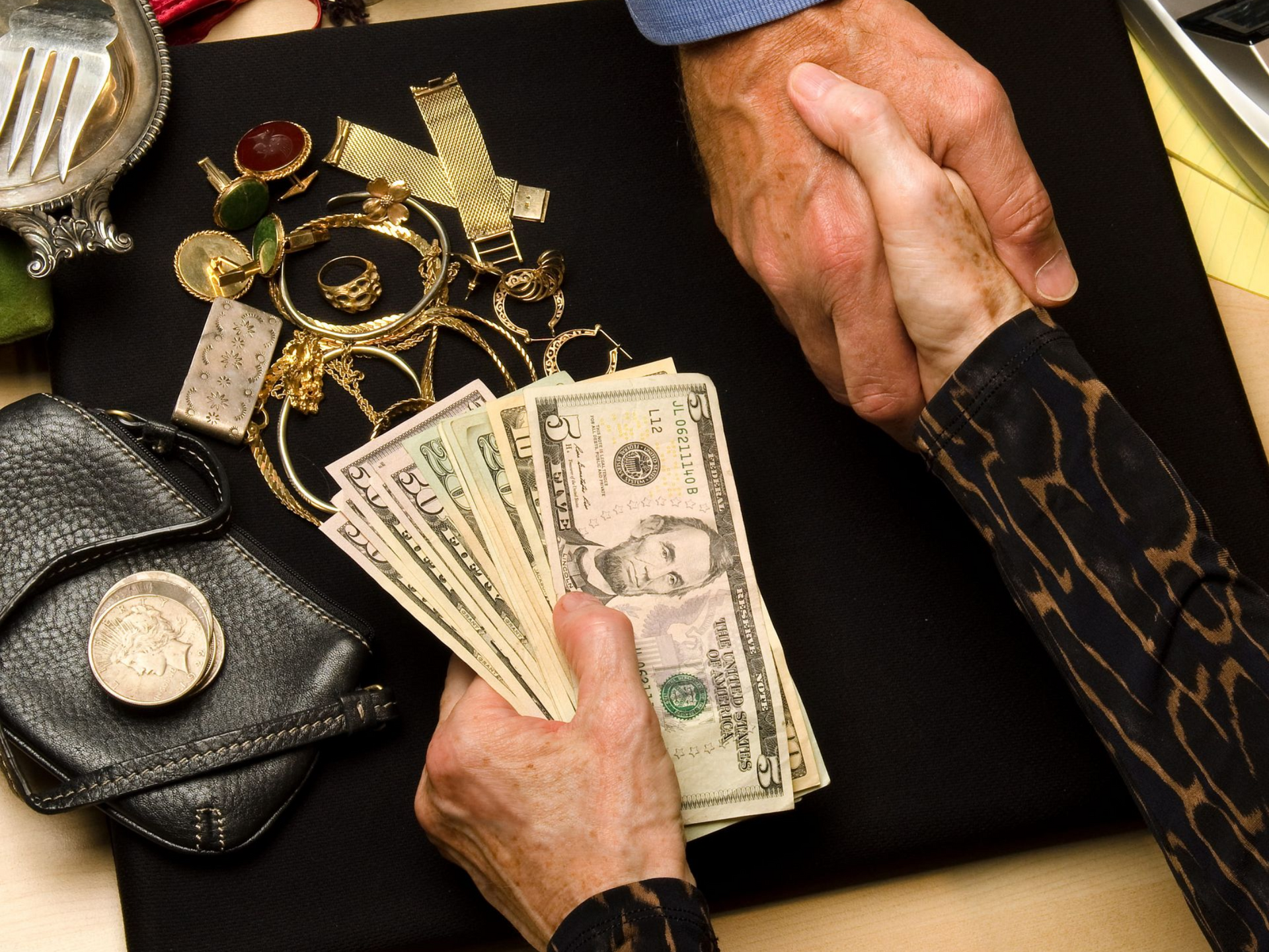 Two people shake hands over a collection of gold jewelry and a fan of US currency laid out on a black surface.