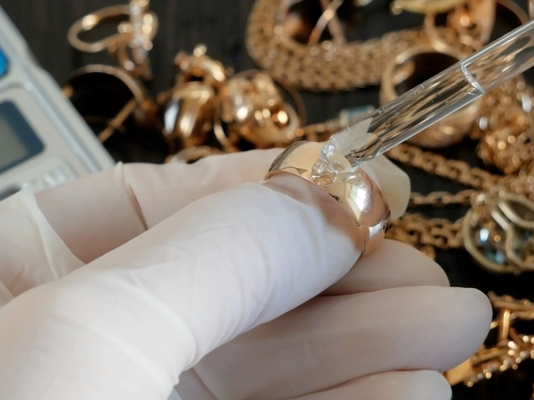 A gloved hand applies a chemical testing drop from a glass dropper onto a gold ring, surrounded by jewelry on a table.