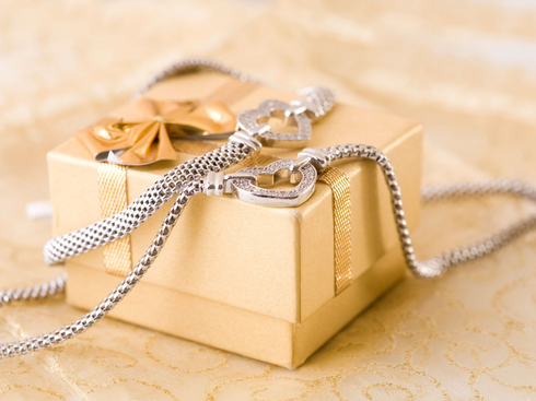 A gold gift box with a ribbon, draped with a delicate silver mesh necklace featuring a heart-shaped clasp.