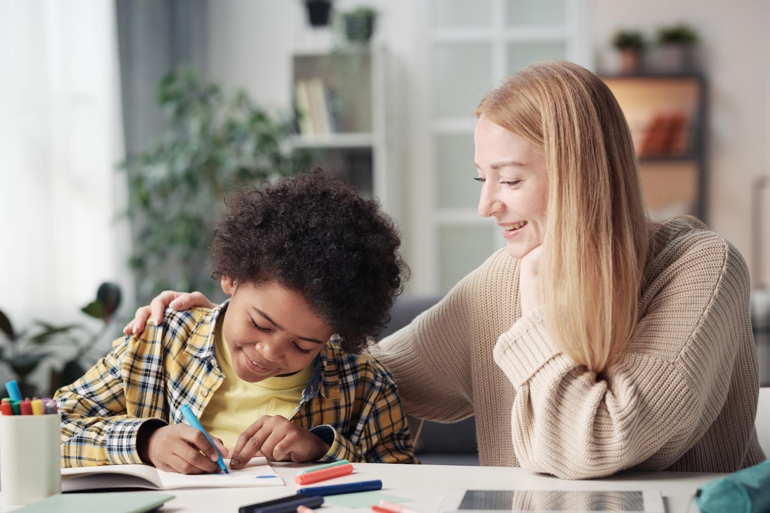 A child, writing at a table, is assisted by a smiling person with a hand on the child's shoulder.
