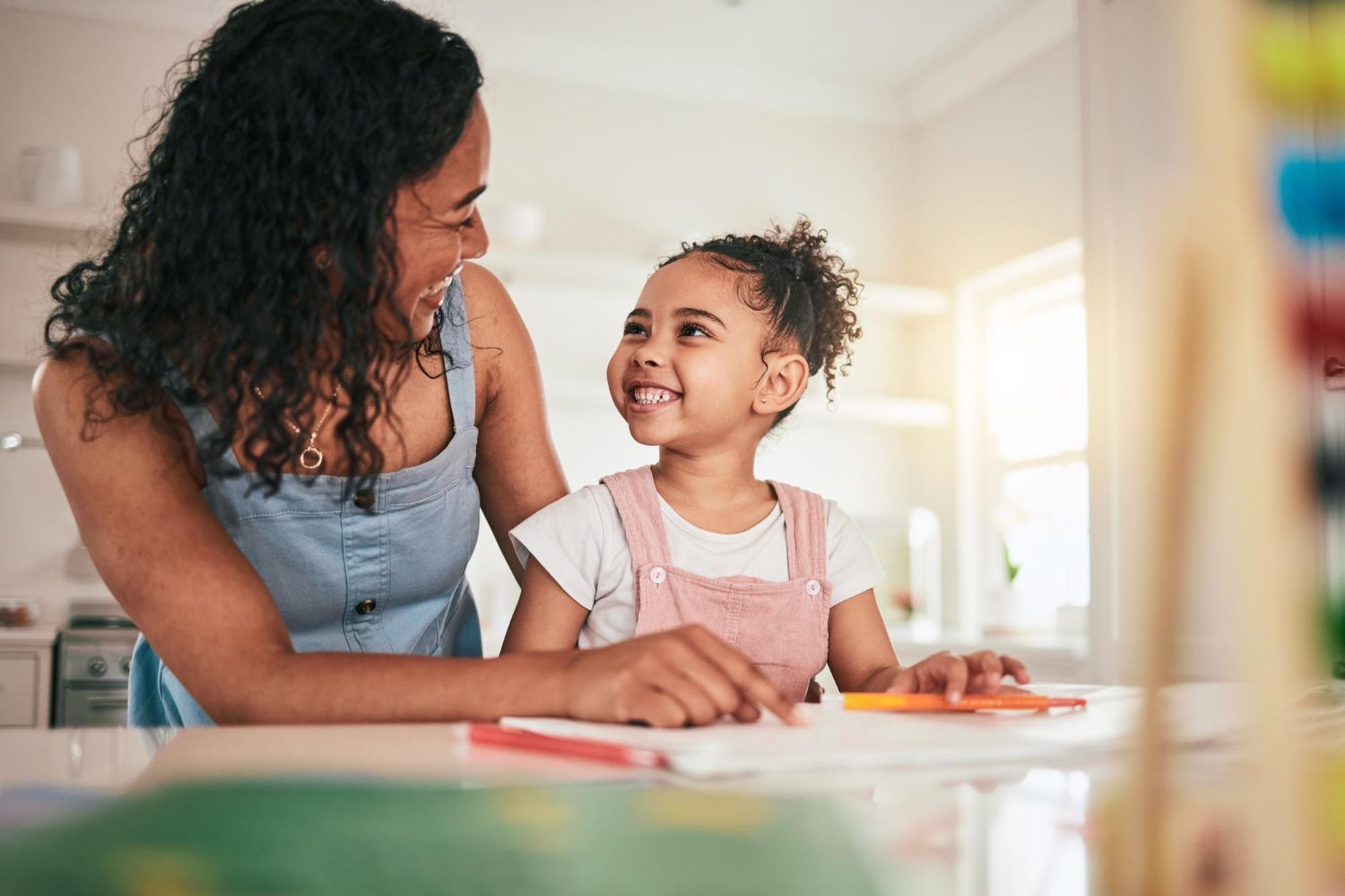 Woman smiling at child drawing at a table, sunny kitchen setting.