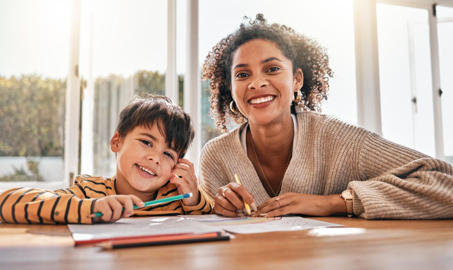 Woman and child smiling, drawing at a table indoors. Light streams through windows.