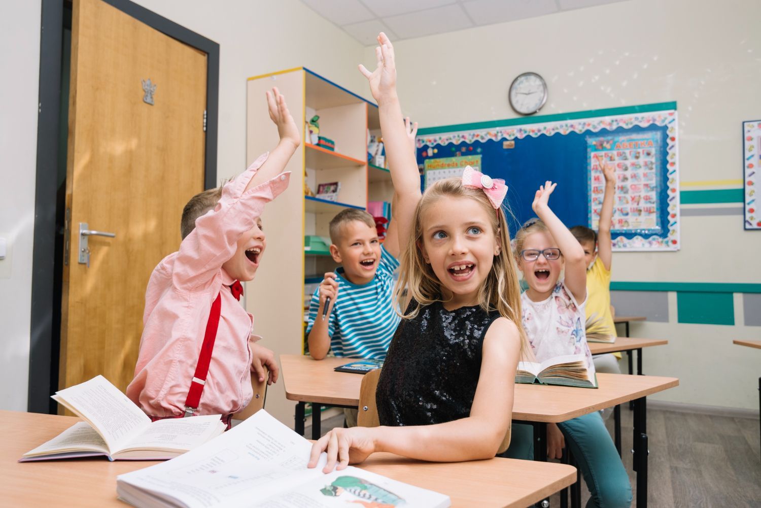 Children in a classroom raise hands enthusiastically, with open books on desks.