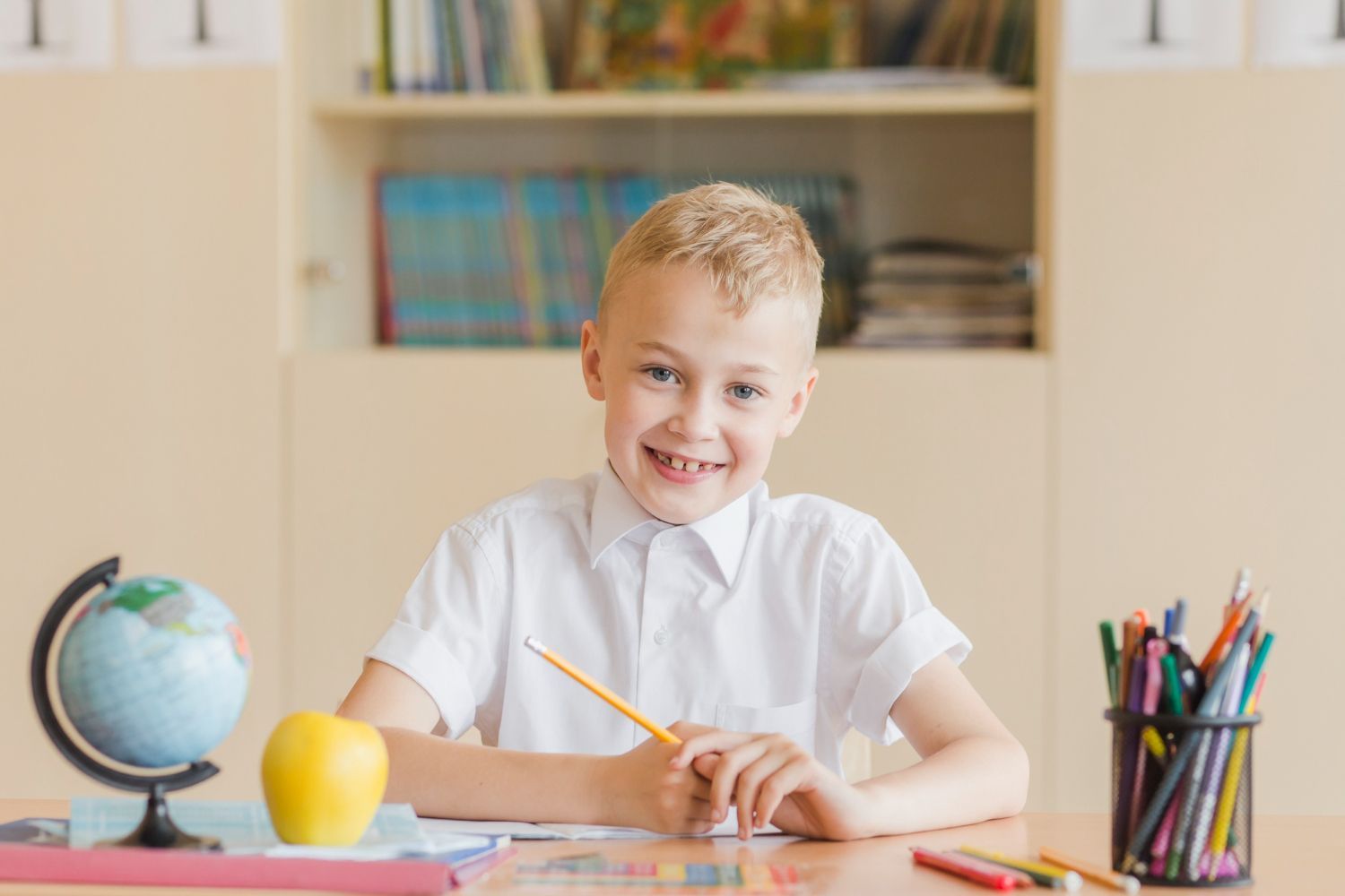 Boy smiling at desk in classroom, holding pencil. Globe, apple, and colored pencils nearby.