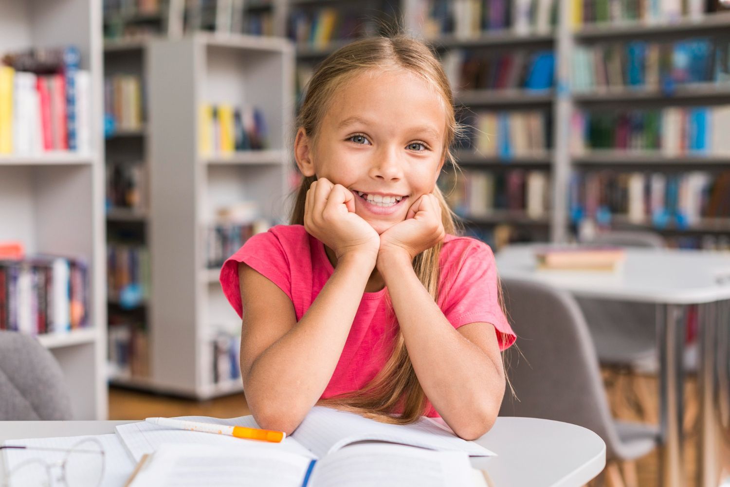 Girl smiling in a library, resting chin on hands, books open on table in front of her. Shelves of books in background.
