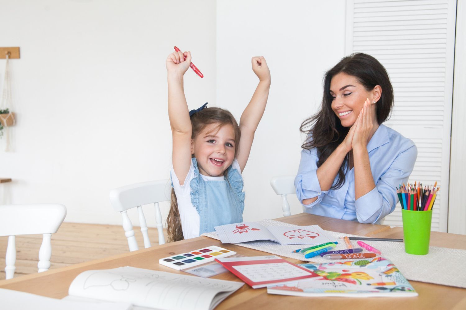 Girl cheering, arms raised, holding crayon; woman smiles, watching. Art supplies on table.