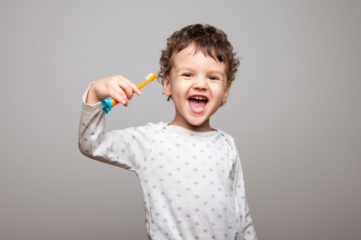Young child with curly hair holding a toothbrush, smiling with open mouth.