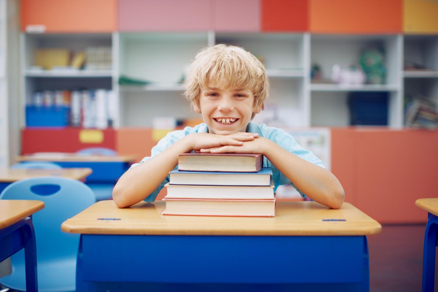Smiling child in a school classroom with books stacked on his desk.