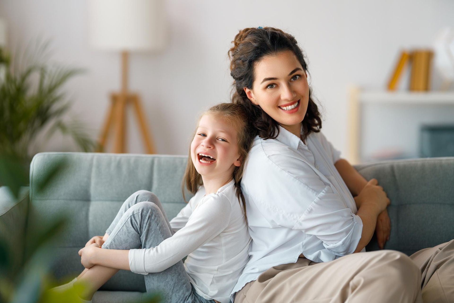 Woman and child sitting back to back on a couch, smiling and laughing. Soft light interior.