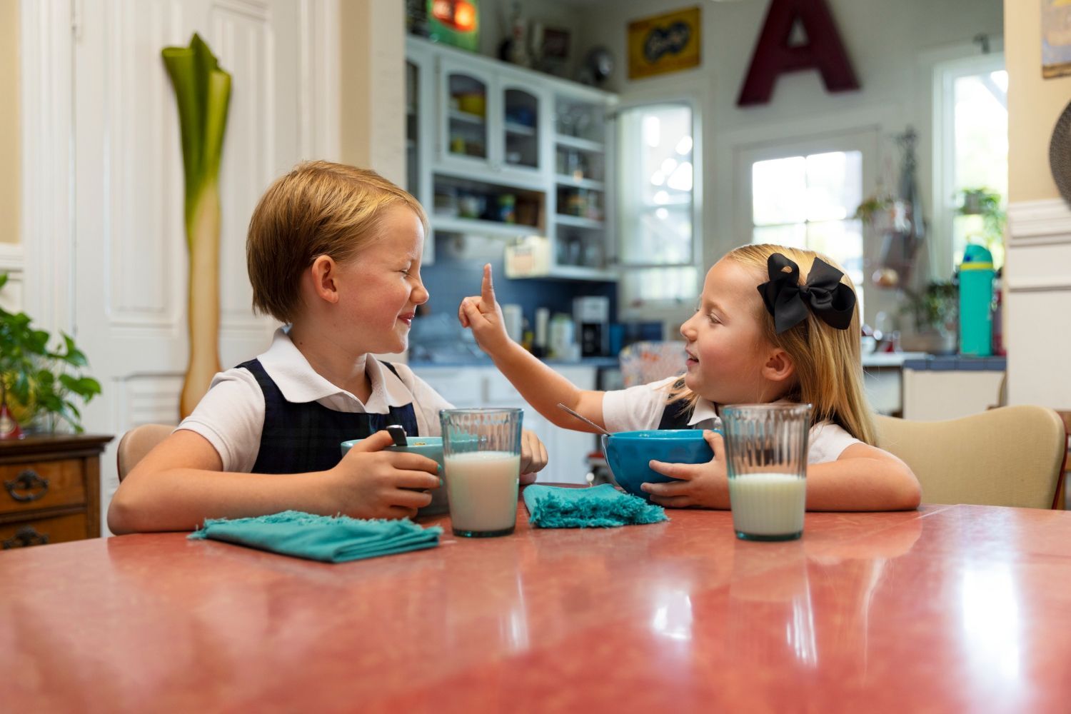 Two children at a table, smiling, having breakfast. Girl points at boy's face. Milk glasses, blue bowls and cloth.