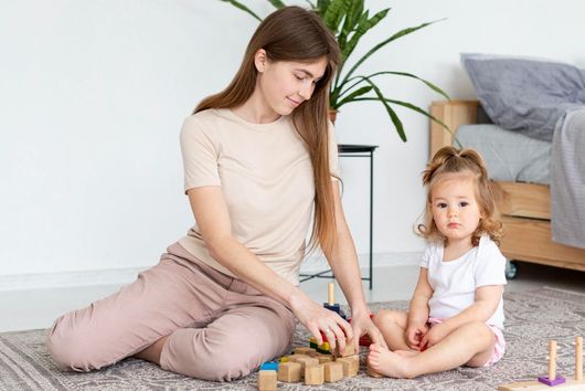 Woman and child playing with wooden blocks on a rug. The child looks at the camera.