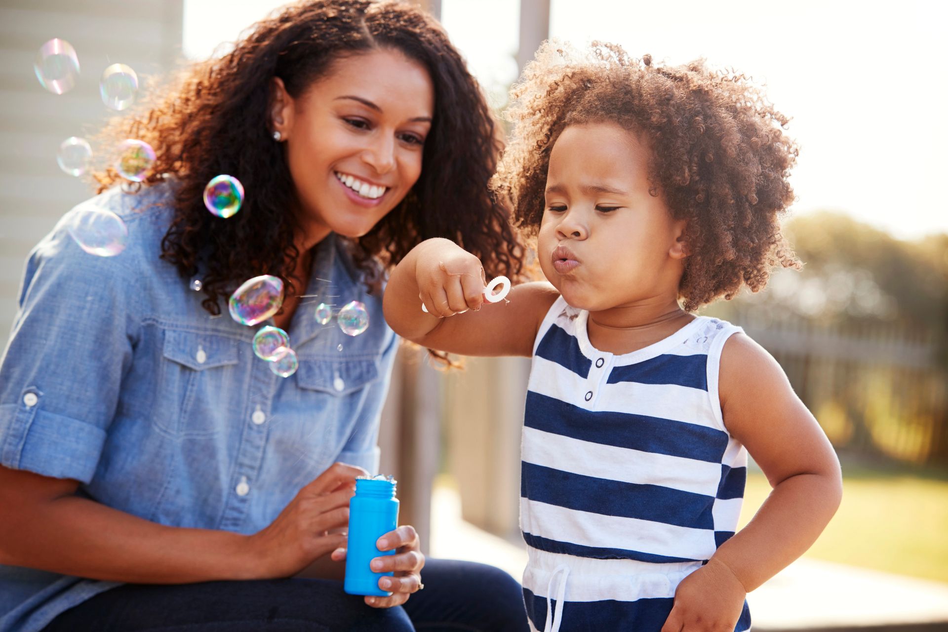 Woman blowing bubbles with a child outdoors; both smiling, child blowing bubbles.