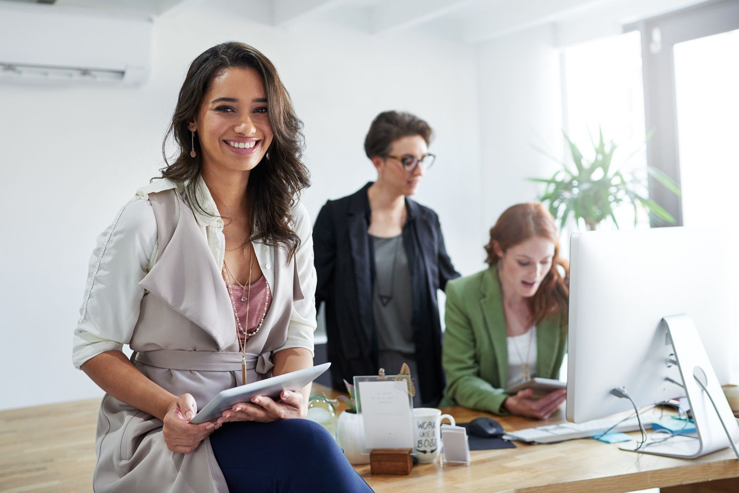Woman smiling, holding tablet, sits on desk in office, two coworkers in background.