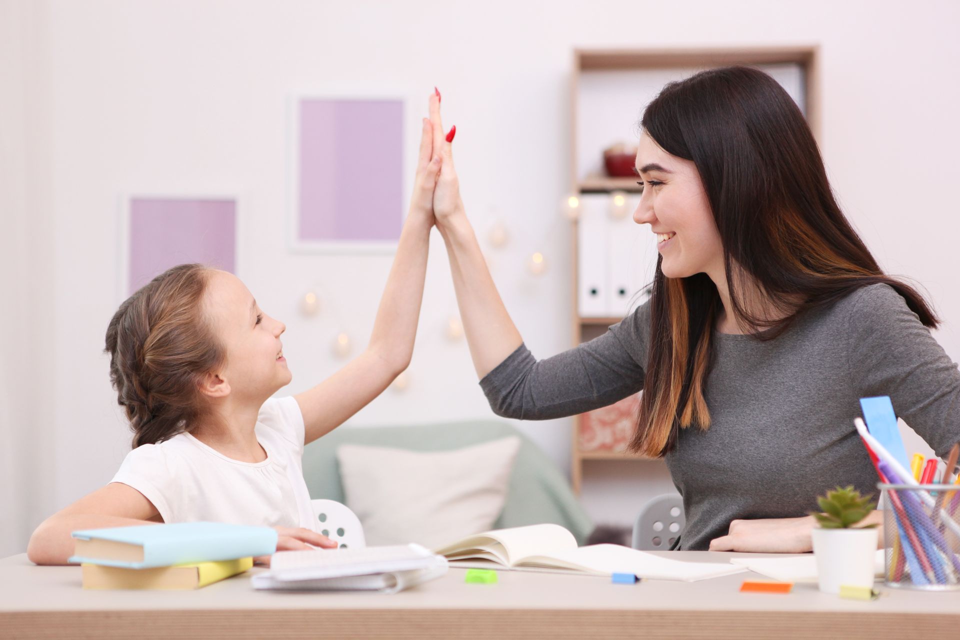 Woman and girl high-fiving at a desk with books and supplies in a home setting.