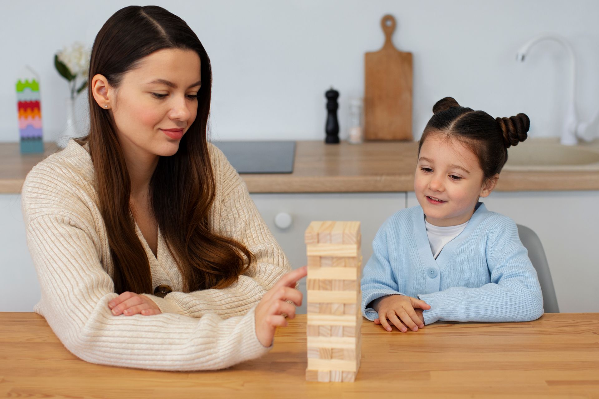 Woman and child playing Jenga at a wooden table in a kitchen; woman reaches for a block, child watches.