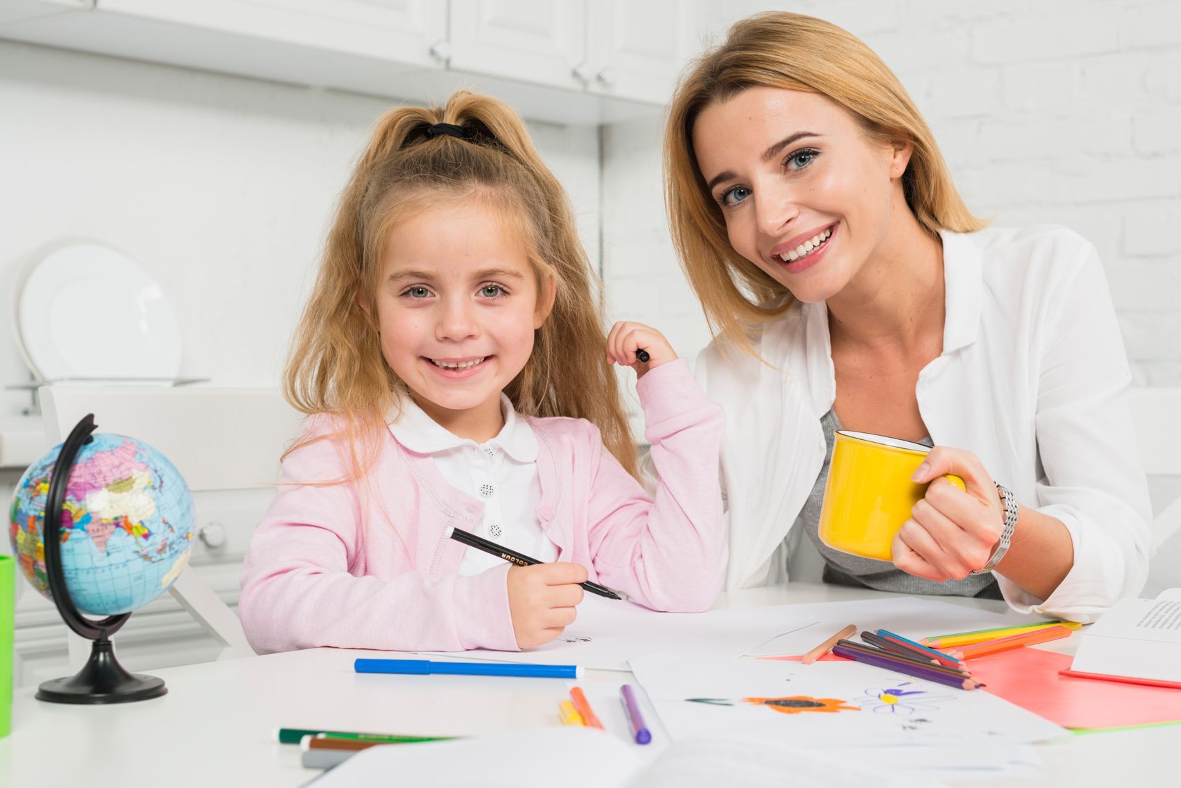 Woman and child smiling, coloring at a table with a globe. Woman holds a yellow mug.