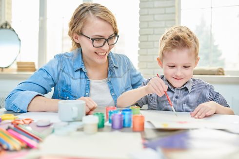 Woman with glasses smiles at a child painting with watercolors at a table with art supplies.