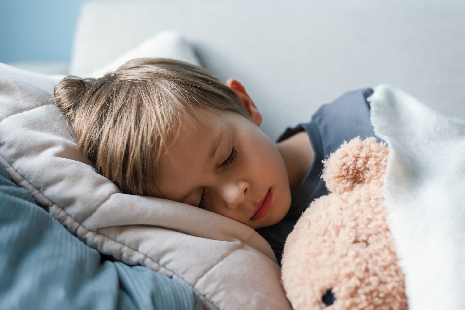 Boy asleep, hugging a teddy bear, under a blanket on a couch.
