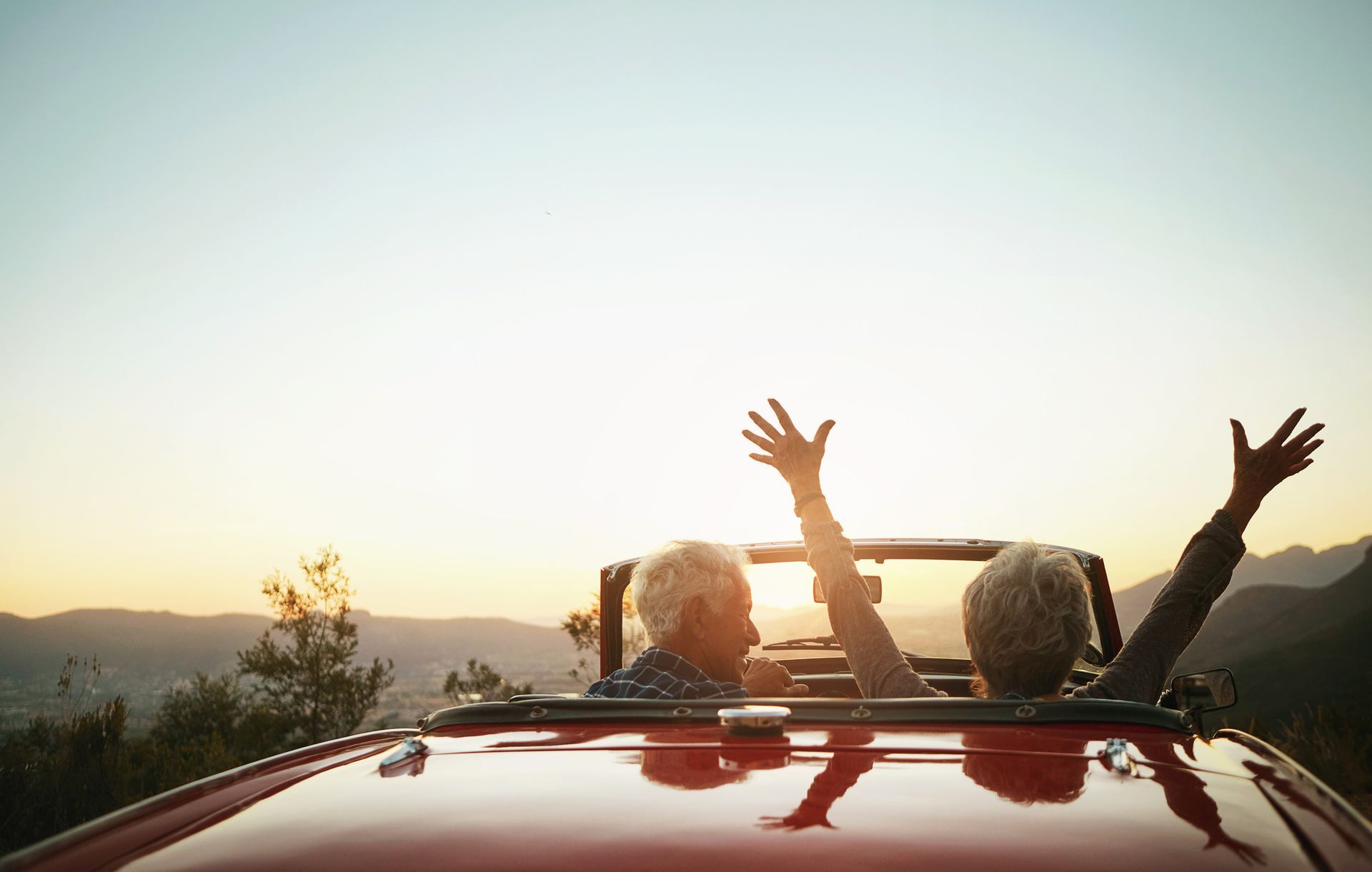 Two people in a red convertible with arms raised at sunset on a scenic road