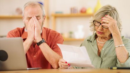 Two stressed people reviewing paperwork at a table, one holding his face and the other rubbing her temple.