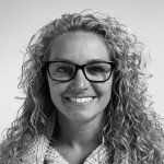 A woman with curly hair and glasses is smiling in a black and white photo.