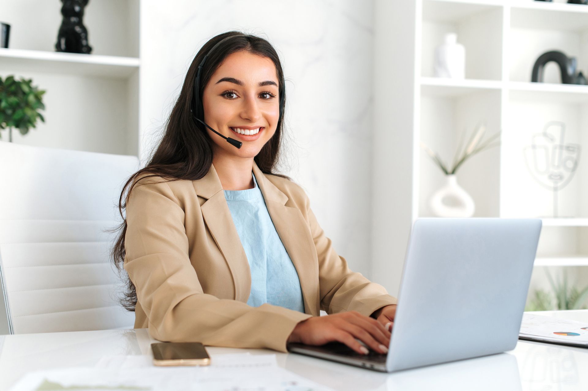 A woman wearing a headset is sitting at a desk using a laptop computer.