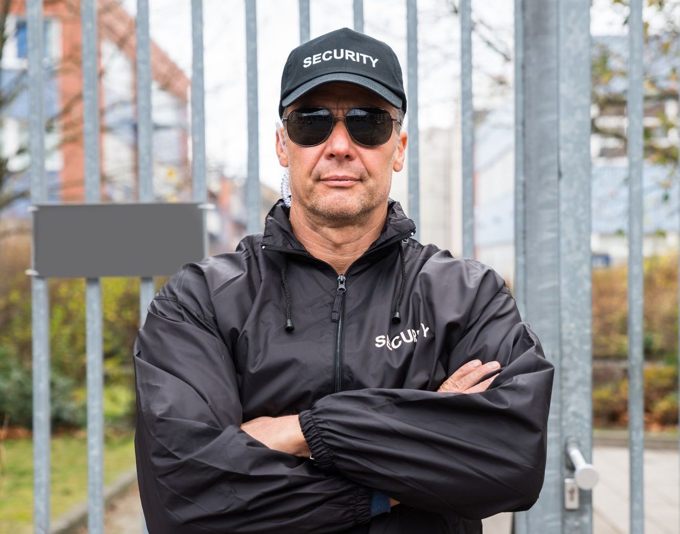 A security guard is standing in front of a fence with his arms crossed.
